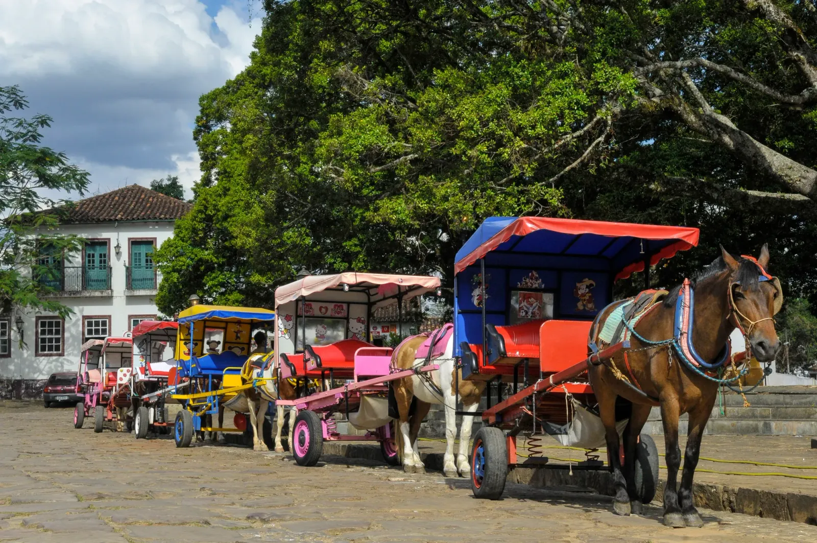 Charretes coloridas paradas na praça de pedra de Tiradentes, à sombra de árvores