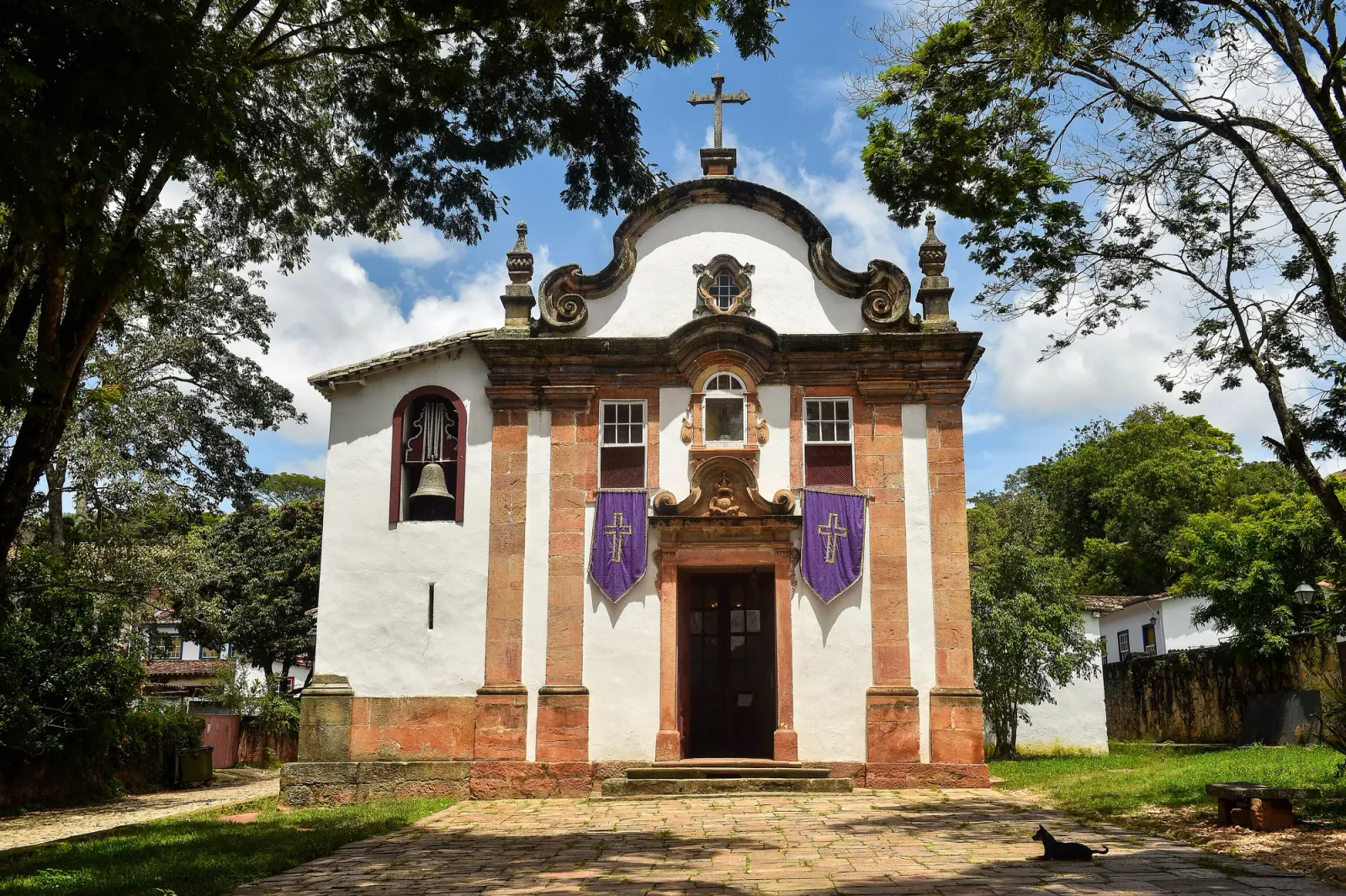 Igreja de Nossa Senhora do Rosário, de fachada branca e portal rosado, entre árvores, em Tiradentes