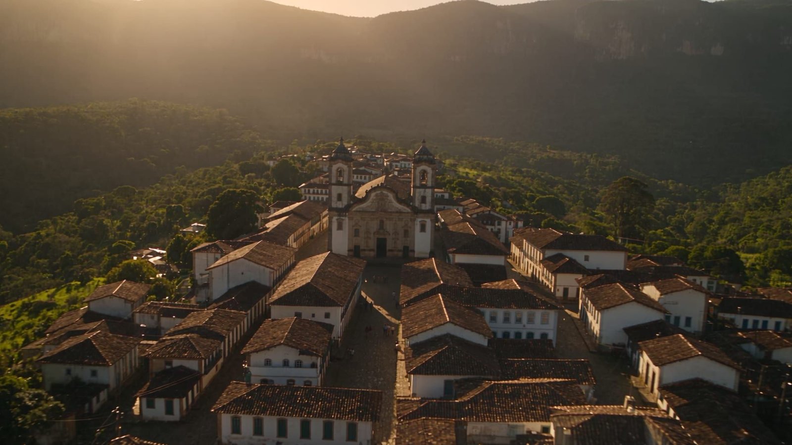 Vista aérea de uma igreja entre casas com telhados de cerâmica, árvores e montanhas ao fundo